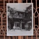 Black-and-white photograph of a two-story balcony house clipped to decorative metal gate